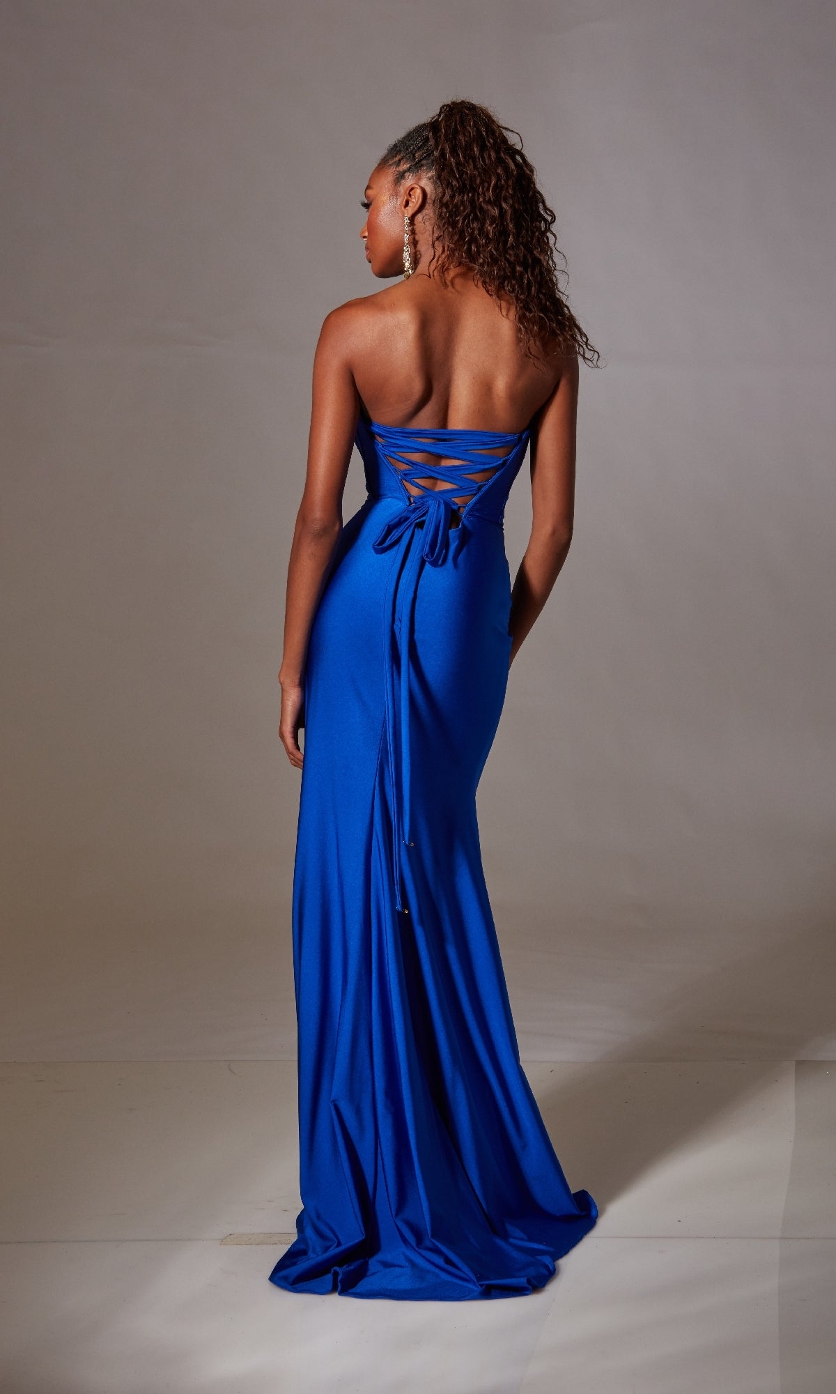 A woman faces away, wearing the Aerie Couture 249 Strapless Long Prom Dress with a corset-style lace-up back and a floor-length blue skirt that flows elegantly against a plain backdrop.